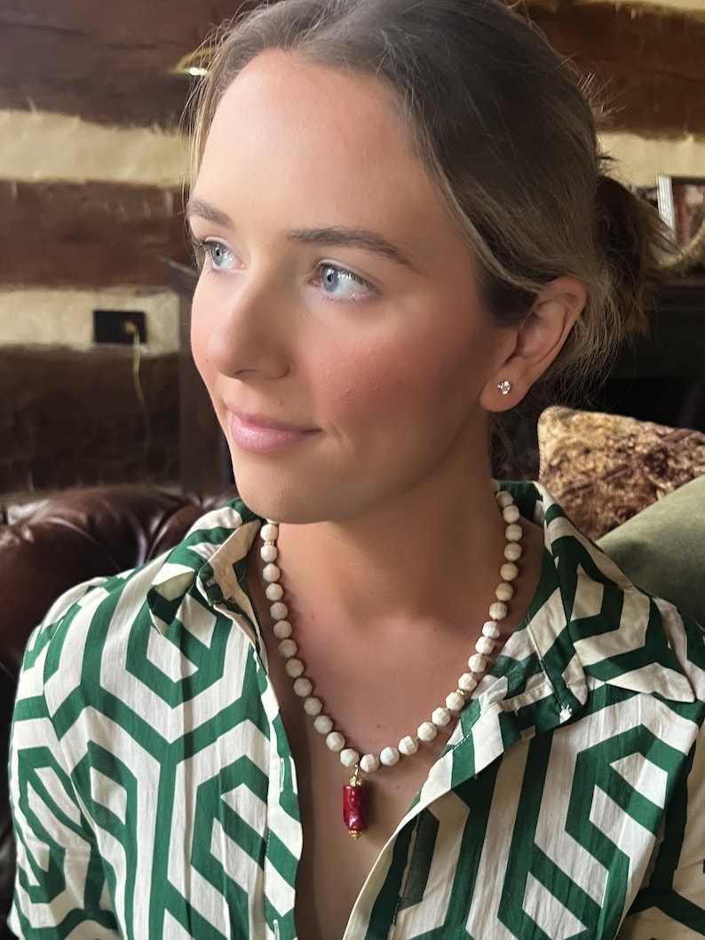 May include: A white and red beaded necklace with a red pendant. The necklace is worn by a person with long brown hair and blue eyes. The person is wearing a green and white patterned shirt.