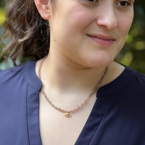 K&ouml;nnte beinhalten: Eine zarte goldene Halskette mit einem kleinen Augen-Anh&auml;nger. Die Kette besteht aus winzigen rosa und klaren Perlen.