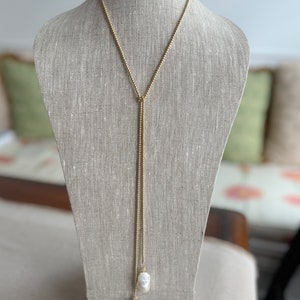 May include: A gold chain necklace with two large white pearls at the end. The necklace is on a mannequin and is styled as a lariat.
