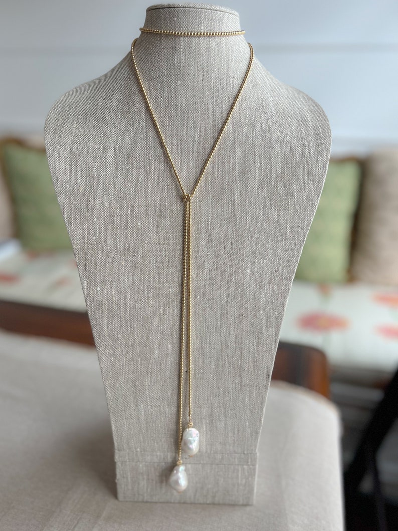 May include: A gold chain necklace with two large white pearls at the end. The necklace is on a mannequin and is styled as a lariat.