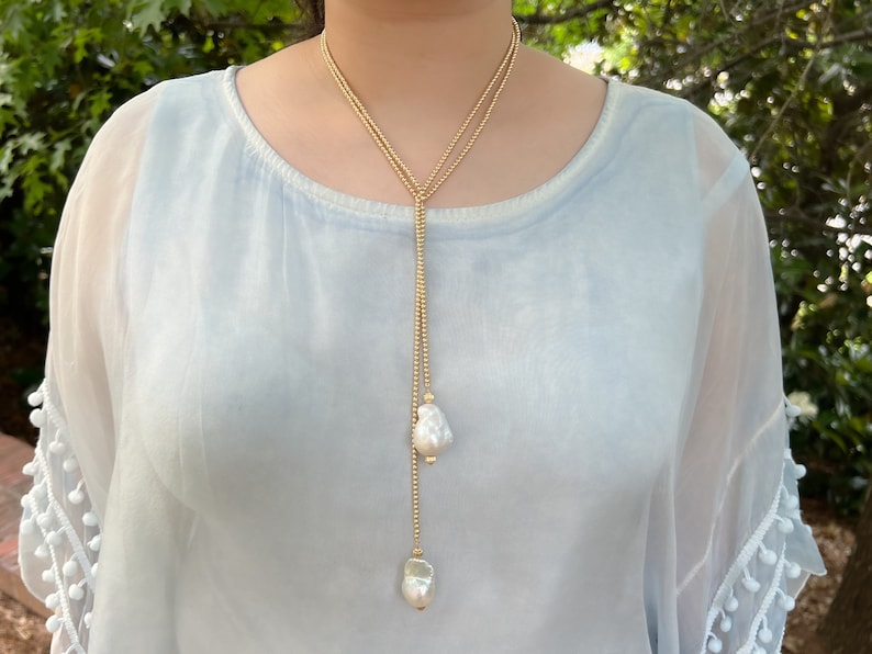 May include: A long gold chain necklace with two large white pearl pendants. The necklace is layered and has a delicate gold chain.