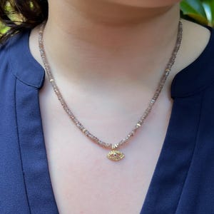 K&ouml;nnte beinhalten: Eine zarte goldene Halskette mit einem kleinen Augen-Anh&auml;nger. Die Halskette besteht aus winzigen braunen Perlen und hat ein goldenes Auge mit einem roten Edelstein in der Mitte.