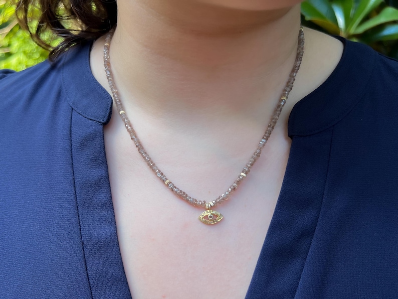 K&ouml;nnte beinhalten: Eine zarte goldene Halskette mit einem kleinen Augen-Anh&auml;nger. Die Halskette besteht aus winzigen braunen Perlen und hat ein goldenes Auge mit einem roten Edelstein in der Mitte.