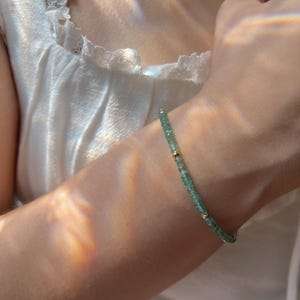 May include: A delicate bracelet featuring small, light green beads strung together with gold-colored accents. The bracelet is worn on a wrist, with a white top in the background. The bracelet is a piece of jewelry.