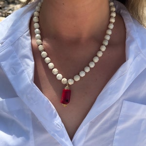 May include: A white beaded necklace with a red coral pendant. The necklace is made of small, round beads and the pendant is a long, thin piece of coral.
