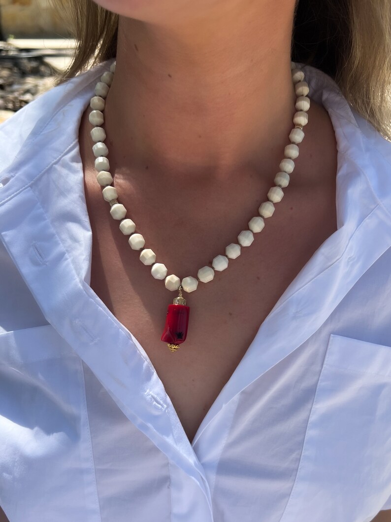 May include: A white beaded necklace with a red coral pendant. The necklace is made of small, round beads and the pendant is a long, thin piece of coral.