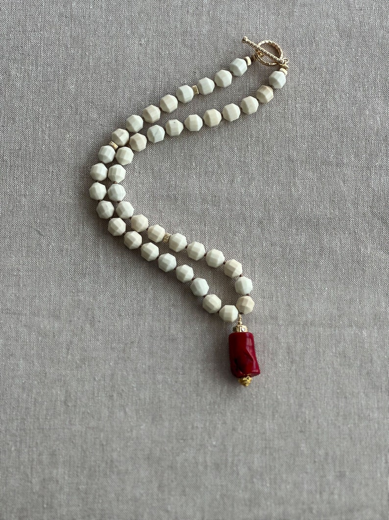 May include: A white ceramic bead necklace with a red coral pendant. The necklace is made of faceted beads and has a gold clasp.