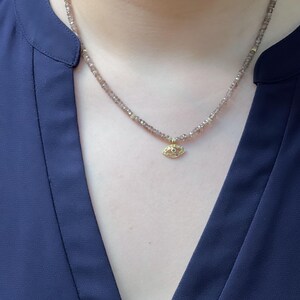K&ouml;nnte beinhalten: Eine zarte Halskette mit einem kleinen goldenen Augen-Anh&auml;nger. Die Halskette besteht aus winzigen braunen Perlen.
