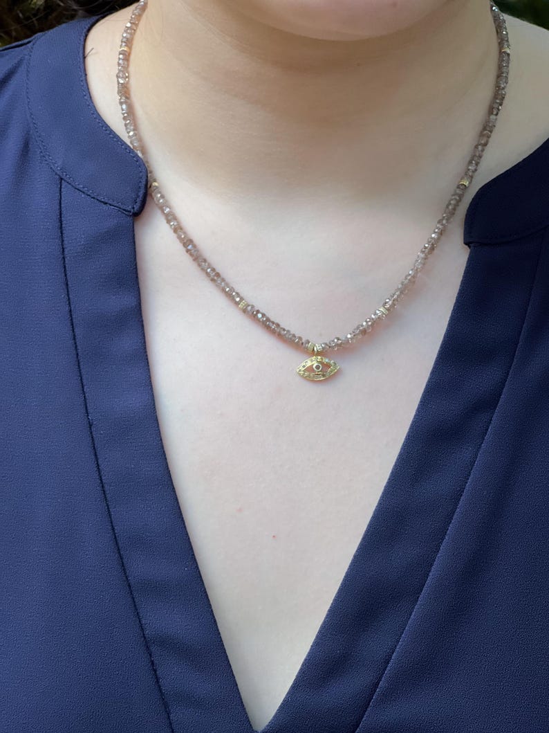 K&ouml;nnte beinhalten: Eine zarte Halskette mit einem kleinen goldenen Augen-Anh&auml;nger. Die Halskette besteht aus winzigen braunen Perlen.