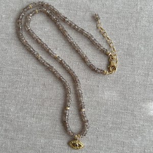 K&ouml;nnte beinhalten: Eine zarte goldene Halskette mit einem kleinen b&ouml;sen Augen-Anh&auml;nger. Die Halskette besteht aus facettierten Rauchquarzperlen und einer goldenen Kette.