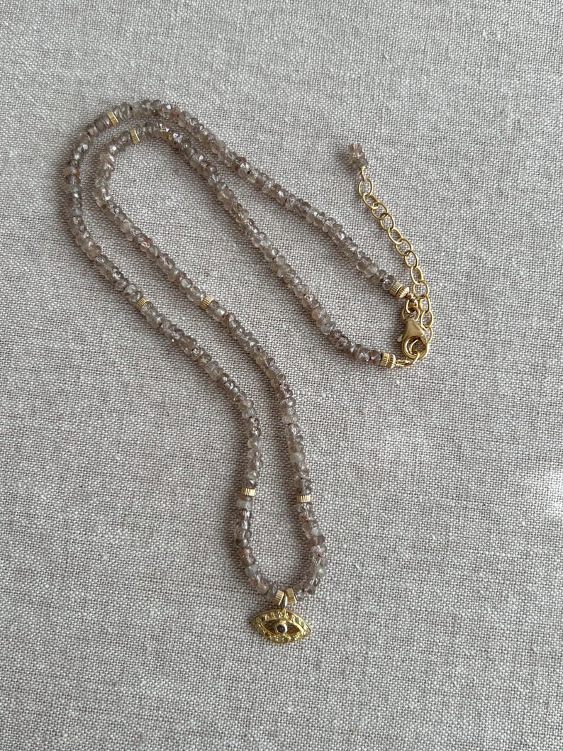 K&ouml;nnte beinhalten: Eine zarte goldene Halskette mit einem kleinen b&ouml;sen Augen-Anh&auml;nger. Die Halskette besteht aus facettierten Rauchquarzperlen und einer goldenen Kette.