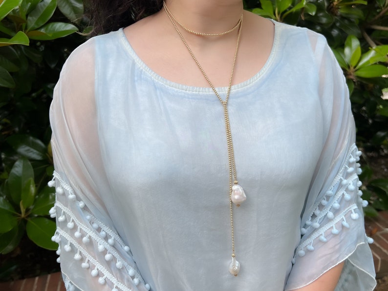 May include: A long gold chain necklace with two large white pearl pendants. The necklace is layered with a shorter gold chain.