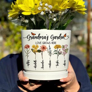 May include: White ceramic flower pot with the text "Grandma's Garden Love Grows Here" and six different flowers with names written below each flower. The names are:  "Ashley", "Elizabeth", "Brandon", "Melonie", "Taylor", and "Olivia".