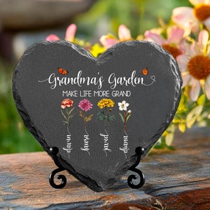 May include: A black heart-shaped slate plaque with the text "Grandma's Garden Make Life More Grand" and five stylized flowers with names written below them. The plaque is on a black metal stand.