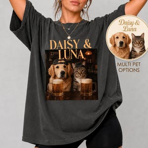 May include: A charcoal gray oversized t-shirt featuring a graphic print of a golden retriever and a tabby cat wearing baseball caps, each sitting with a glass of beer. The text Daisy & Luna appears above the image, with a circular badge showing the pets.
