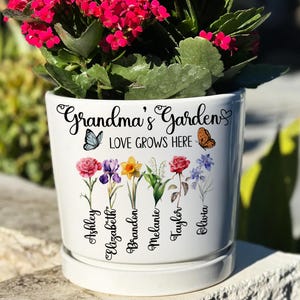 May include: White ceramic Grandma's Garden flower pot with a saucer, featuring the text "Grandma's Garden LOVE GROWS HERE" and floral illustrations. Names Ashley, Elizabeth, Brandon, Melanie, Taylor, and Olivia are printed below the flowers. The pot is filled with a plant with red flowers.