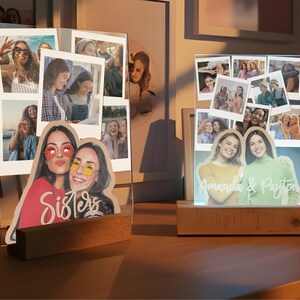 May include: Two custom acrylic photo displays with wooden bases. The left display features a collage of photos and a cutout of two people with the word Sisters. The right display shows a photo collage and a cutout of two people with the text Amanda & Payton.