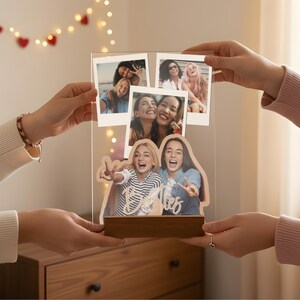 May include: A clear acrylic photo display board featuring three polaroid-style snapshots at the top and a larger custom-cut photo of two people at the bottom. The word Besties is written in white script across the bottom photo, all set on a wooden base.