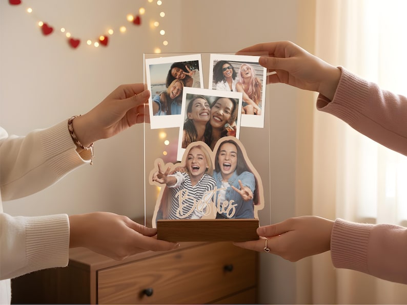 May include: A clear acrylic photo display board featuring three polaroid-style snapshots at the top and a larger custom-cut photo of two people at the bottom. The word Besties is written in white script across the bottom photo, all set on a wooden base.