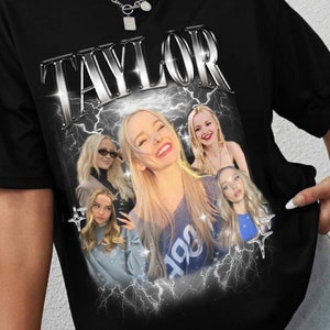 May include: Black t-shirt with a graphic print of a young woman with blonde hair. The graphic includes the word "TAYLOR" in silver letters with lightning bolts behind the text and images.