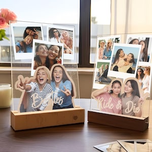 May include: Two clear acrylic photo displays with wooden bases. The left display features a cutout of two people with the text Besties. The right display features a cutout of two people with the text Ride or Die. Both include multiple photo prints.