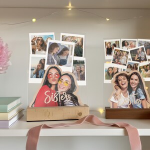 May include: Two clear acrylic photo displays featuring multiple polaroid-style snapshots and a custom cutout of two people at the base. The left display has a light wood base with the word Sisters, and the right has a dark wood base with Besties.