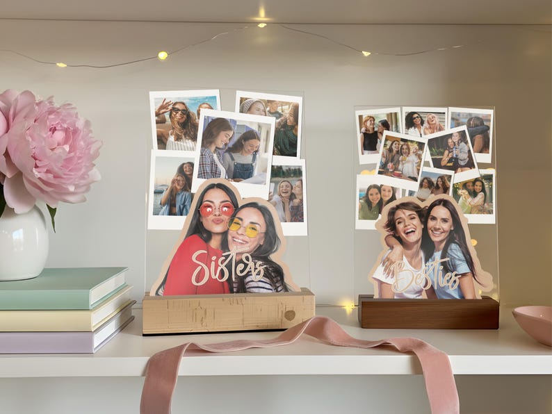 May include: Two clear acrylic photo displays featuring multiple polaroid-style snapshots and a custom cutout of two people at the base. The left display has a light wood base with the word Sisters, and the right has a dark wood base with Besties.