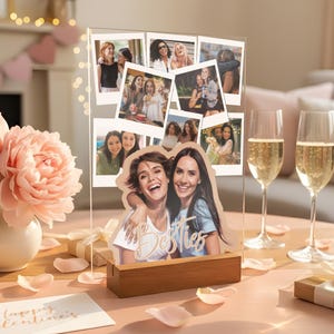 May include: A clear acrylic photo display stand with a wooden base featuring a collage of various candid photographs and a larger cutout image of two smiling individuals with the word Besties written in white script across the front surface.