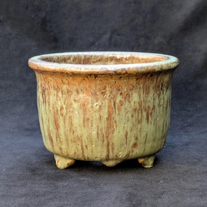 Pode incluir: Um pequeno vaso redondo de cerâmica com uma abertura larga e três pés pequenos. O vaso tem um esmalte manchado verde e castanho, com uma superfície texturizada. A borda é de cor castanha escura.
