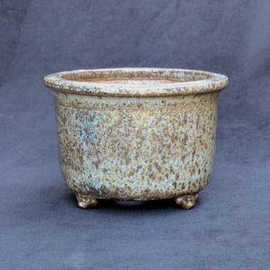 May include: A small, round ceramic planter with a speckled, earthy brown and blue glaze. The planter has a slightly flared rim and sits on three small feet. This decorative pot is suitable for small plants and succulents.