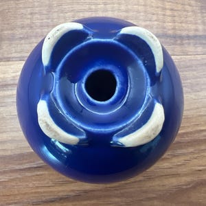 May include: A cobalt blue ceramic vase with a glossy finish. The vase has a circular shape with a central opening and four white, petal-like accents around the base. The vase is set against a wooden surface.