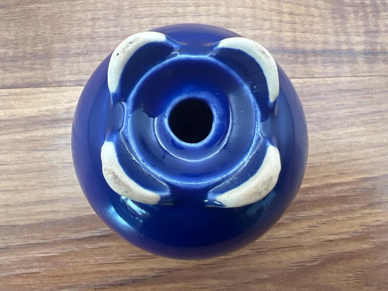 May include: A cobalt blue ceramic vase with a glossy finish. The vase has a circular shape with a central opening and four white, petal-like accents around the base. The vase is set against a wooden surface.