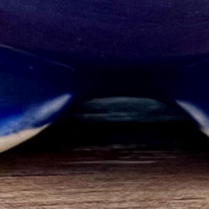May include: A close-up of a ceramic bowl with a deep blue glaze. The bowl has three small, rounded feet with a white rim. The feet are visible at the bottom of the image, resting on a wooden surface.