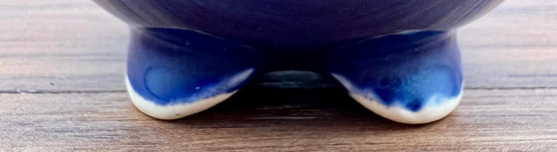 May include: A close-up of a ceramic bowl with a deep blue glaze. The bowl has three small, rounded feet with a white rim. The feet are visible at the bottom of the image, resting on a wooden surface.