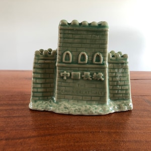 May include: A green ceramic figurine of the Great Wall of China. The figurine has three towers and the text "The China Great Wall" is written on the front.