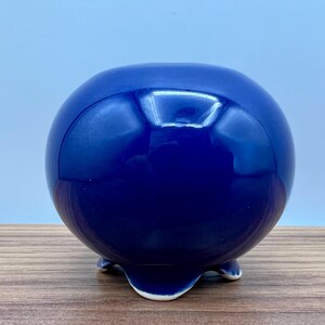May include: A glossy, spherical, cobalt blue ceramic planter with three small feet. The planter has a smooth, reflective surface and a rounded shape, suitable for small plants or decorative use.
