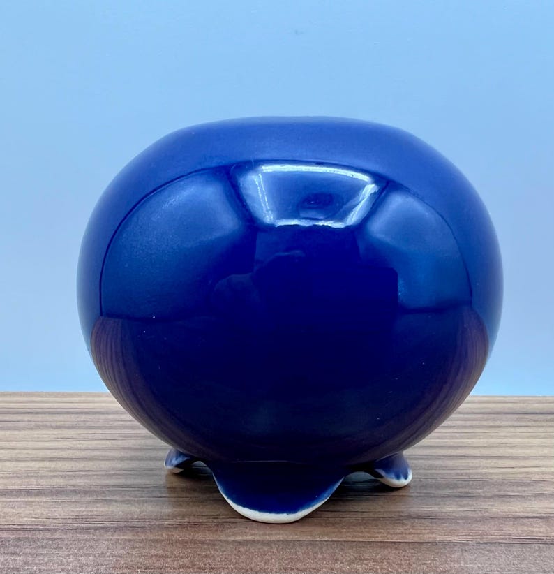 May include: A glossy, spherical, cobalt blue ceramic planter with three small feet. The planter has a smooth, reflective surface and a rounded shape, suitable for small plants or decorative use.