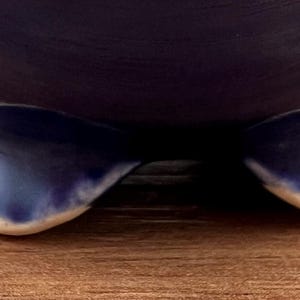 May include: A dark blue ceramic bowl with a rounded shape, resting on three curved feet. The feet are a lighter shade of blue with a white trim. The bowl sits on a wooden surface.