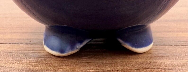 May include: A dark blue ceramic bowl with a rounded shape, resting on three curved feet. The feet are a lighter shade of blue with a white trim. The bowl sits on a wooden surface.