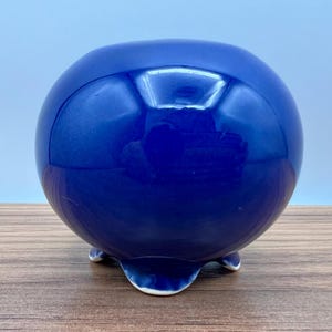 May include: A round, cobalt blue ceramic bowl with a glossy finish. The bowl rests on three small, white-tipped feet. The spherical shape and deep blue color give it a modern, minimalist aesthetic. This decorative piece could be used as a planter or a decorative accent.