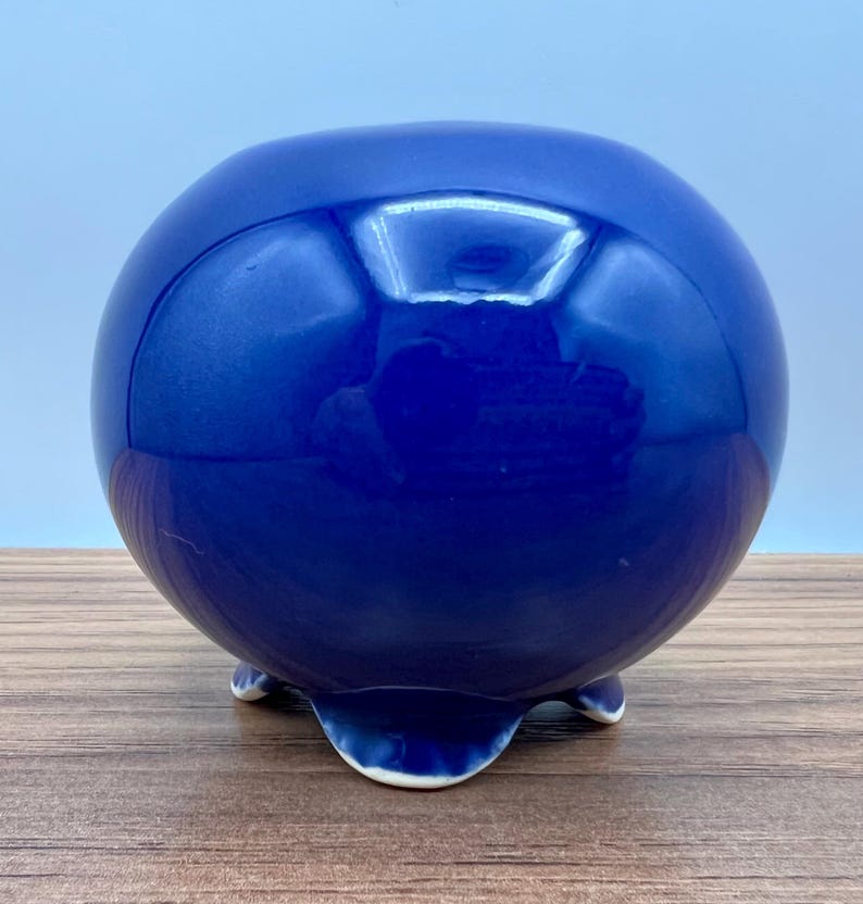 May include: A round, cobalt blue ceramic bowl with a glossy finish. The bowl rests on three small, white-tipped feet. The spherical shape and deep blue color give it a modern, minimalist aesthetic. This decorative piece could be used as a planter or a decorative accent.