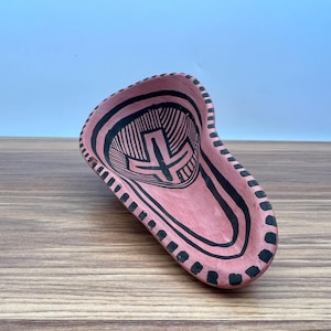 May include: A hand-painted, pink ceramic dish with a unique, elongated shape. The dish features a black geometric design on the interior and around the rim. The design includes lines, shapes, and a central cross-like symbol.