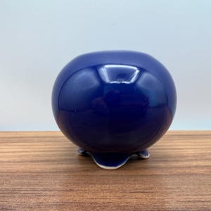 May include: A glossy, spherical, navy blue ceramic pot with three small feet. The pot has a smooth, reflective surface and sits on a wooden surface. The design is simple and elegant, suitable for small plants or decorative use.
