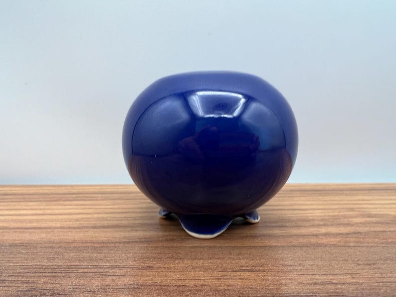 May include: A glossy, spherical, navy blue ceramic pot with three small feet. The pot has a smooth, reflective surface and sits on a wooden surface. The design is simple and elegant, suitable for small plants or decorative use.
