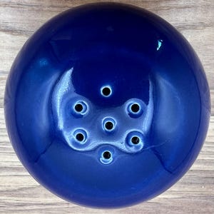 May include: A deep blue ceramic flower frog with a glossy finish. The frog has a rounded shape with multiple holes for arranging flower stems. The holes are arranged in a circular pattern. The flower frog is designed to hold flowers in place.