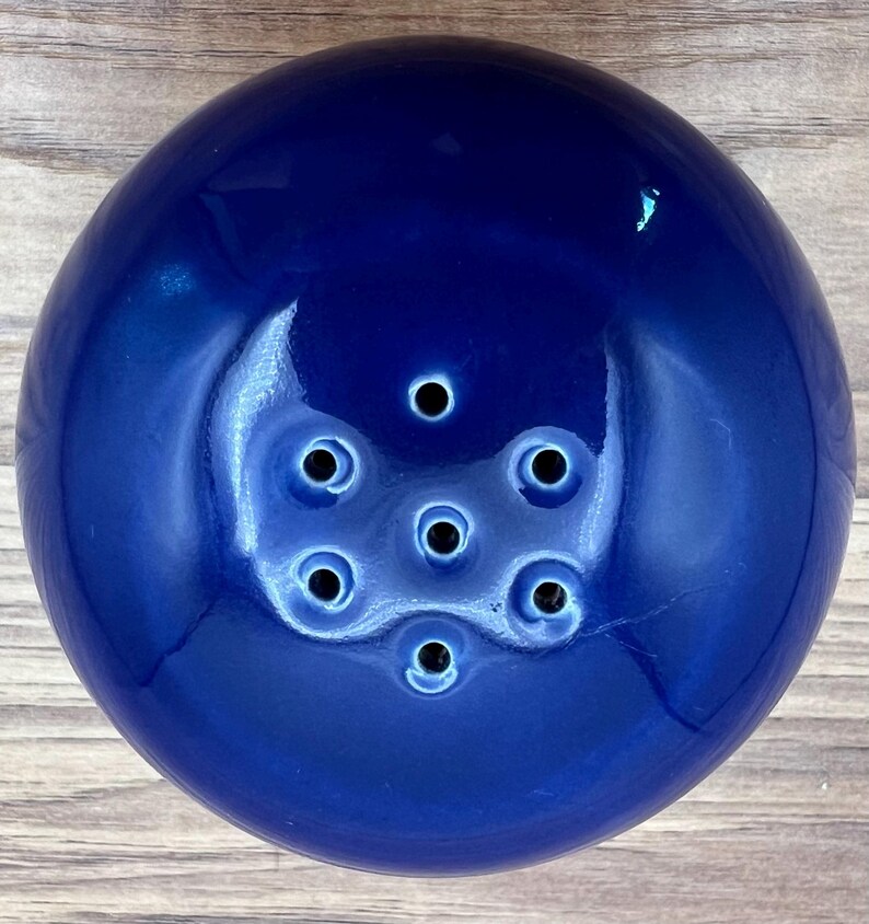 May include: A deep blue ceramic flower frog with a glossy finish. The frog has a rounded shape with multiple holes for arranging flower stems. The holes are arranged in a circular pattern. The flower frog is designed to hold flowers in place.