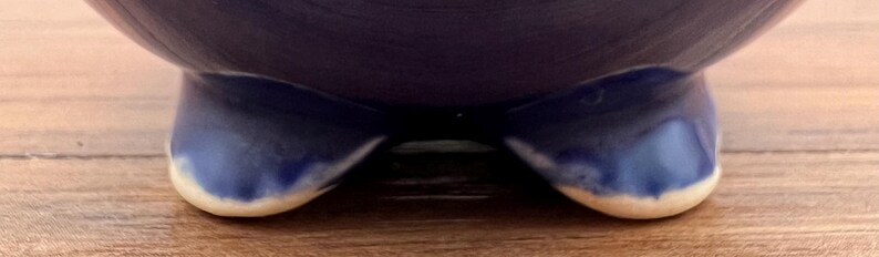 May include: A dark blue ceramic bowl with three rounded feet. The feet have a lighter blue gradient with a white edge. The bowl sits on a light brown wooden surface.