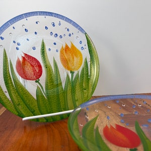 May include: Two clear glass plates with a blue rim and a white background. The plates have a painted design of green grass, red and yellow tulips, and blue dots.