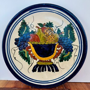 May include: A white ceramic plate with a blue rim and a hand-painted design of a fruit bowl with grapes, pears, and a pineapple. The bowl is painted in shades of blue, yellow, green, and red.