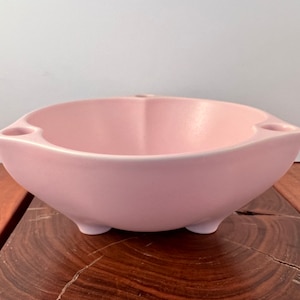 May include: A light pink ceramic bowl with three small holes around the rim and three small feet on the bottom.